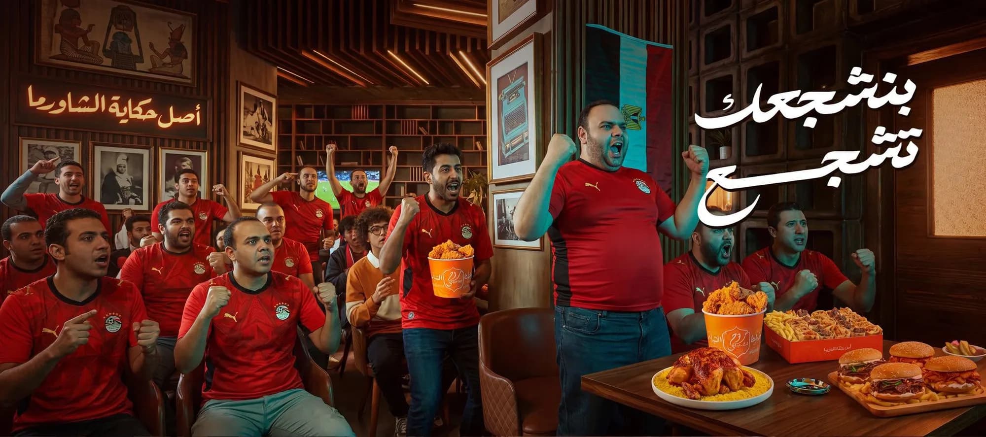 مجموعة من المشجعين بقمصان المنتخب المصري يشاهدون مباراة في مطعم روح دمشق بطنطا مع وجبات دجاج ومشاوي.