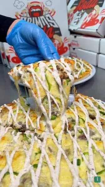 شريحة بيتزا من بريموز بيتزا في مارينا العلمين مغطاة بصوص أبيض وجبنة تمط، مع علب البيتزا في الخلفية.