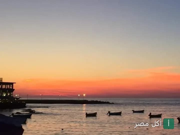 منظر الغروب على البحر بجانب مطعم SeaSide في الإسكندرية مع قوارب صغيرة في المياه.