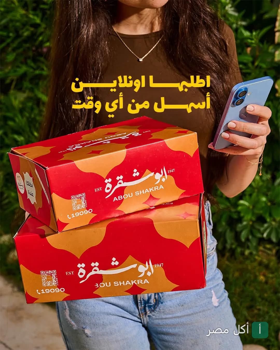 شخص يحمل علب تيك أواي حمراء خاصة بمطعم أبو شقرة مع هاتف محمول لطلب الأوردر أونلاين.