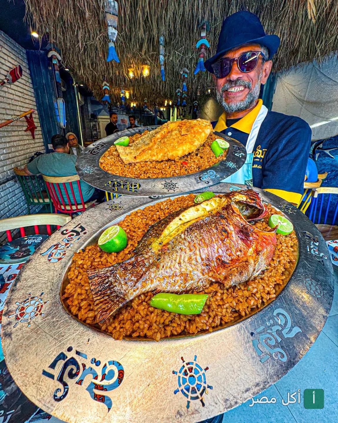 موظف في مطعم بهيج يقدم أطباق سمك مشوي ومقلي مع أرز الصيادية في ديكور بحري.