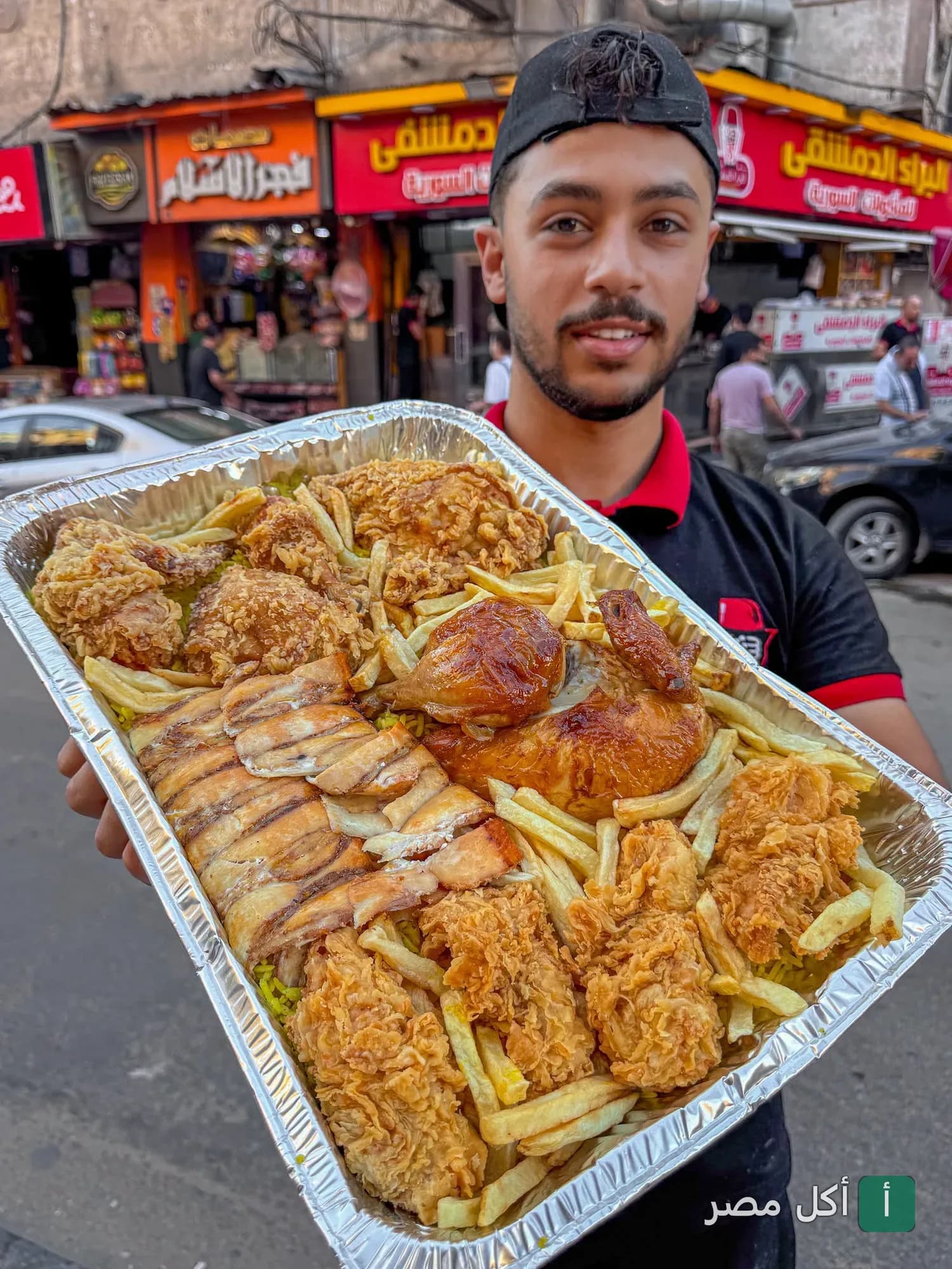 موظف في مطعم البراء الدمشقي 2 بالعامرية يحمل صينية كبيرة تحتوي على دجاج مقلي ومشاوي وبطاطس محمرة.