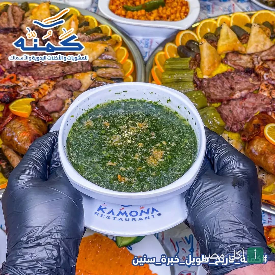 طبق ملوخية طازجة يقدمه مطعم كمنه للمشويات في سيدي جابر مع صواني مشاوي وأكلات بدوية في الخلفية.