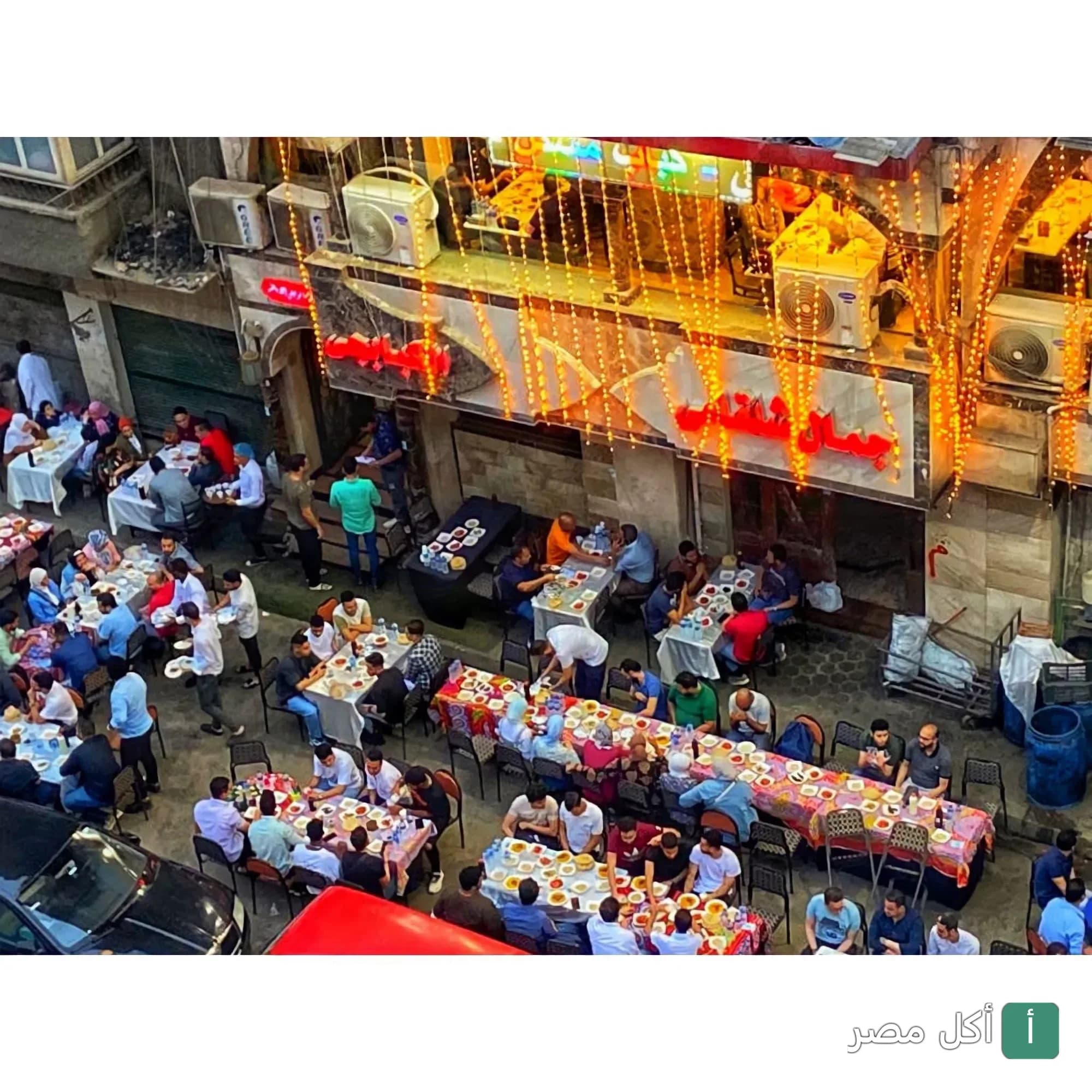 لقطة علوية لمطعم جمال شلقامى الكبابجى في الجيزة، يظهر الزبائن يتناولون المشويات في طاولات خارجية مضاءة بالأنوار.