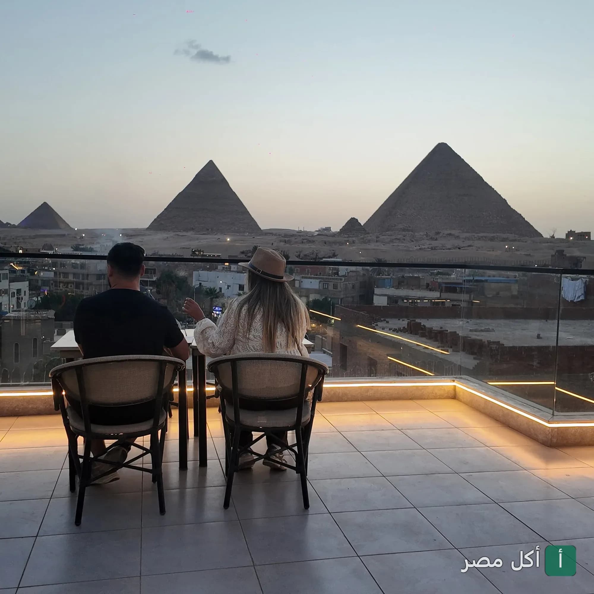 زوجان يجلسان في تراس 139 بيراميدز لاونج بنزلة السمان مع إطلالة مباشرة على أهرامات الجيزة وقت الغروب.