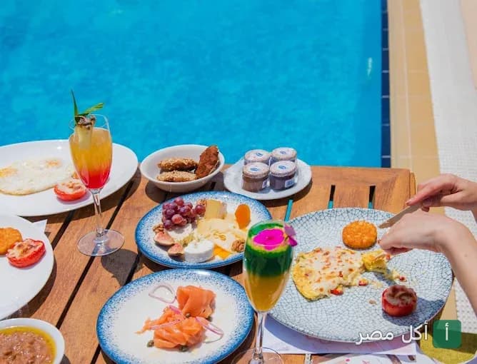 فطور متنوع مع مشروبات ملونة على طاولة خشبية بجانب حمام السباحة في مطعم ذا رووف توب بجاردن سيتي.