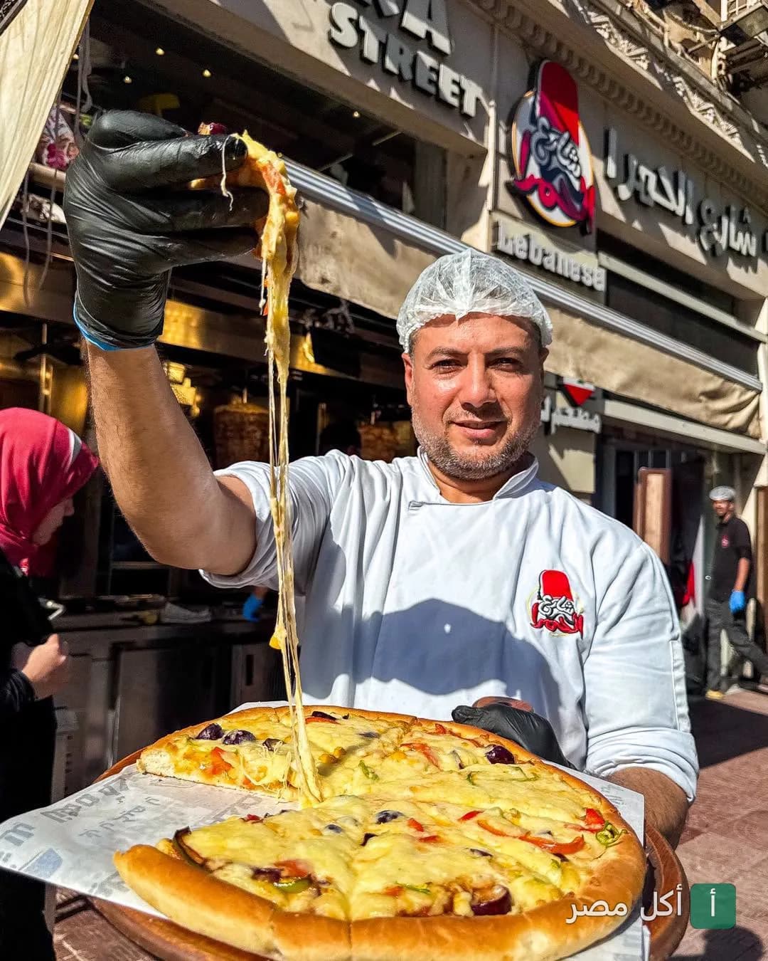 شيف في مطعم شارع الحمرا بباب اللوق يرفع قطعة بيتزا بالجبنة السايحة أمام واجهة المطعم.