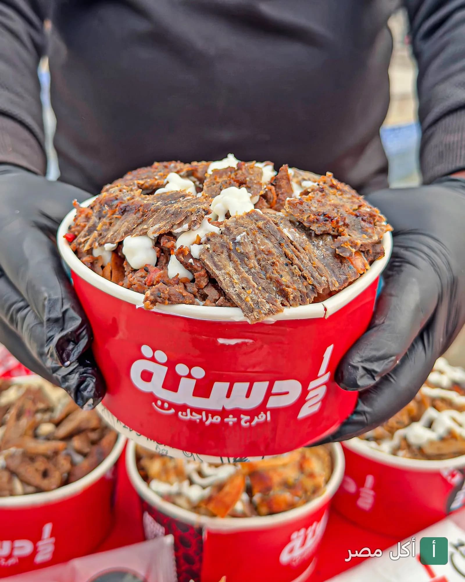 شخص يرتدي قفازات سوداء يحمل علبة حمراء من مطعم نص دستة مليئة بشرائح الشاورما المصرية مع صوص أبيض.
