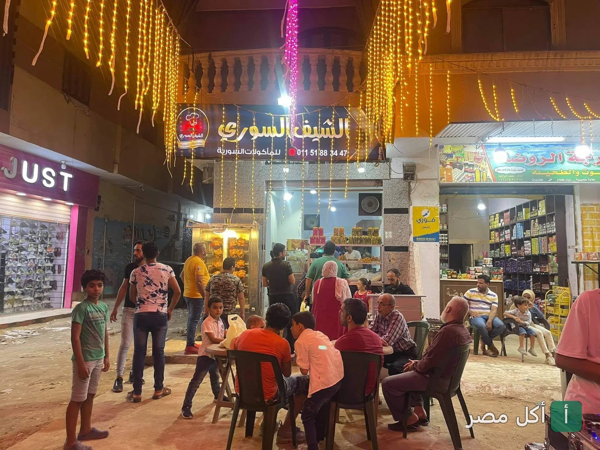 واجهة مطعم الشيف السوري في 6 أكتوبر ليلاً مع زبائن يجلسون في الخارج وزينة إضاءة معلقة.