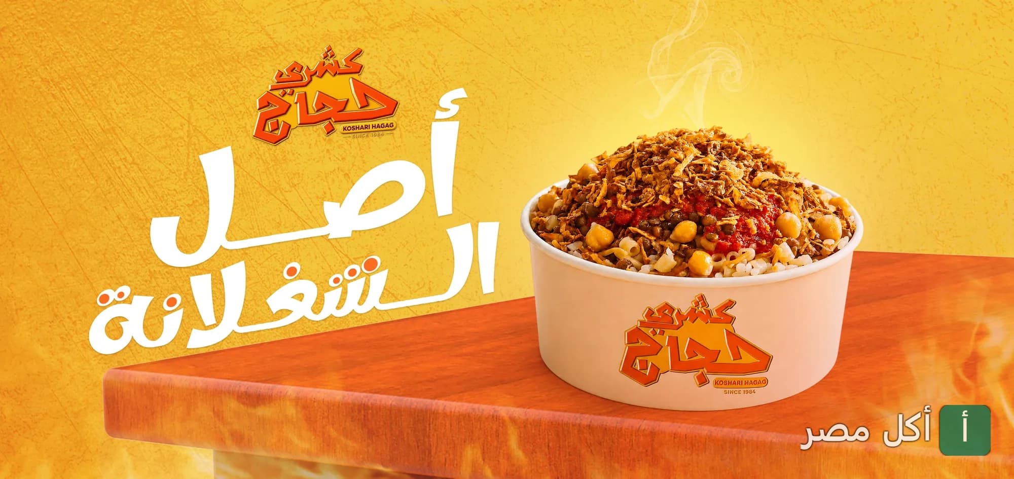 طبق كشري ساخن في علبة مطعم كشري حجاج بإمبابة مع شعار المطعم وخلفية صفراء.