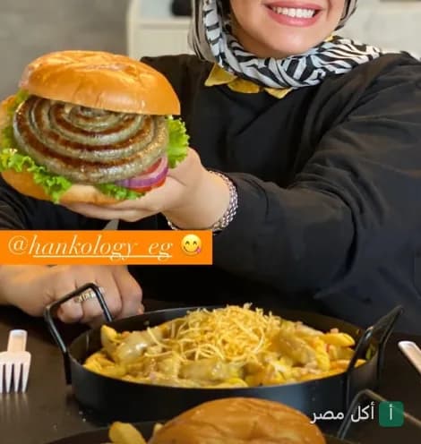 شخص يمسك ساندوتش برجر سجق حلزوني في مطعم هانكولوجي بالشيخ زايد مع طبق بطاطس بالجبنة.