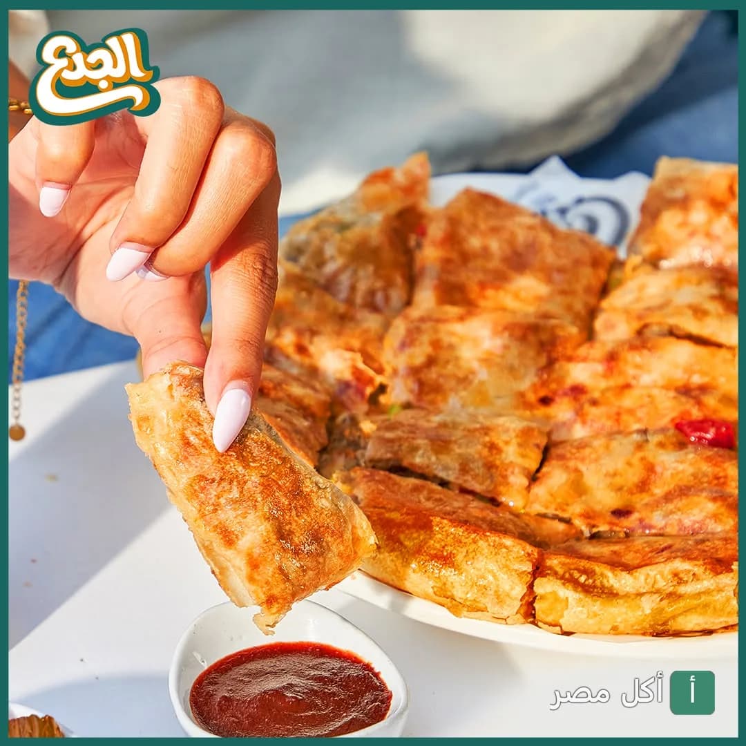 شخص يمسك قطعة من فطير شرقي محشو من مطعم الجدع في الشيخ زايد مع صوص أحمر.