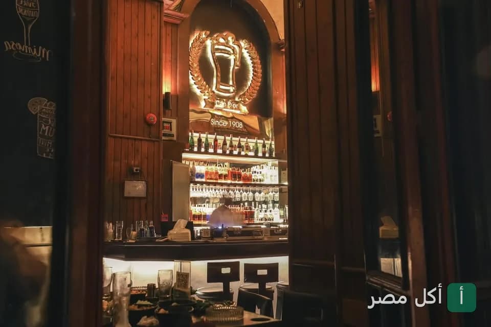 واجهة البار الخشبي الكلاسيكي في مطعم كاب دور بعابدين مع إضاءة دافئة وشعار المطعم التاريخي.