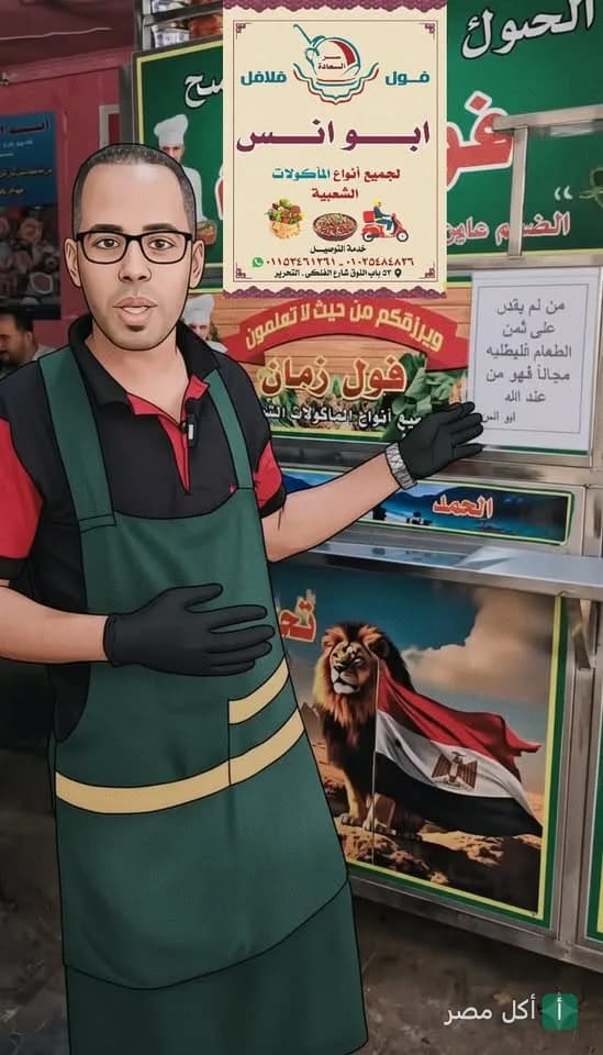 موظف يرتدي مئزرًا أخضر يشير إلى لافتة مطعم فول ابو انس في عابدين.