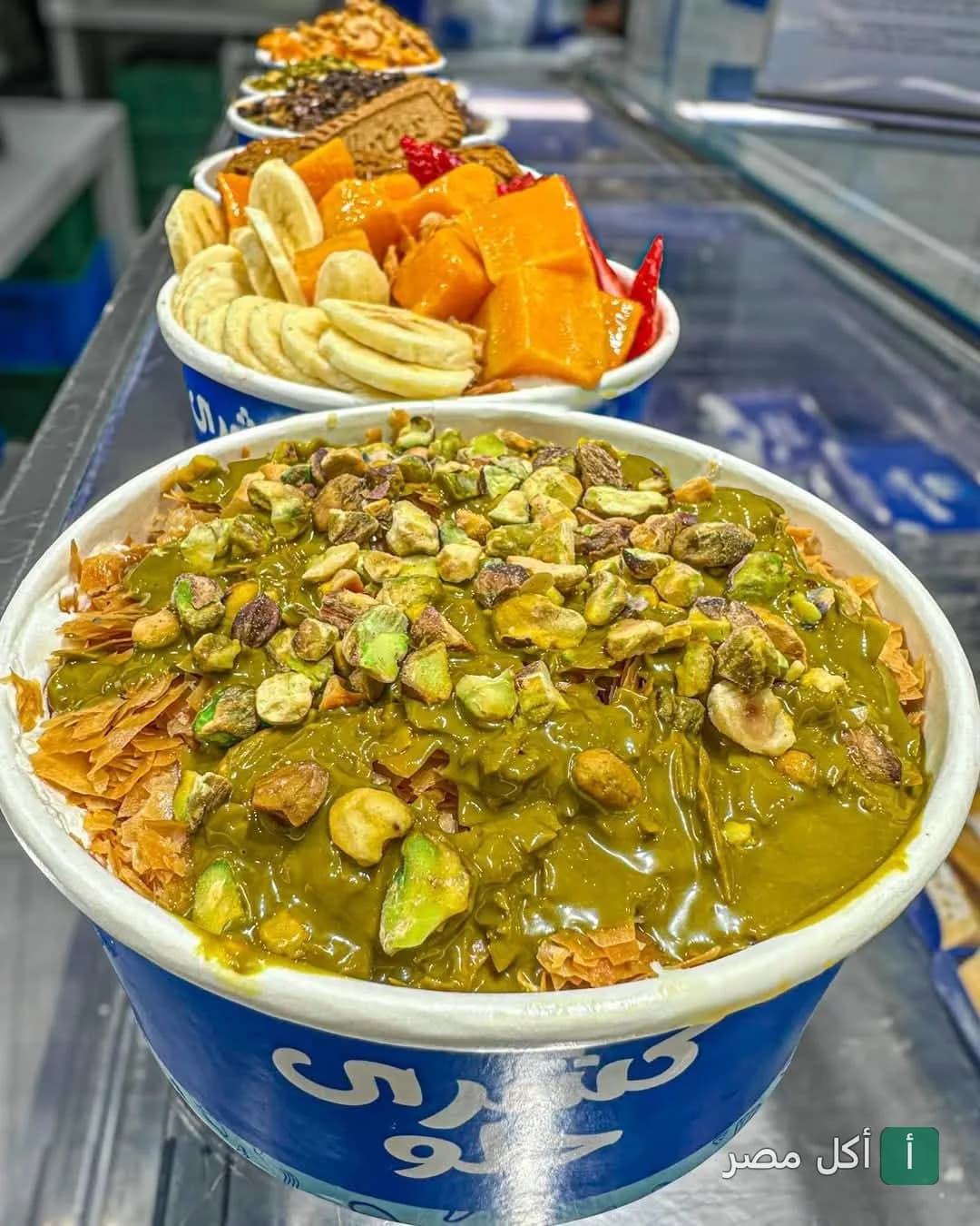 علب حلويات بـ لبن في عين شمس، تظهر واحدة مغطاة بصوص الفستق والمكسرات والأخرى بقطع المانجو والموز.