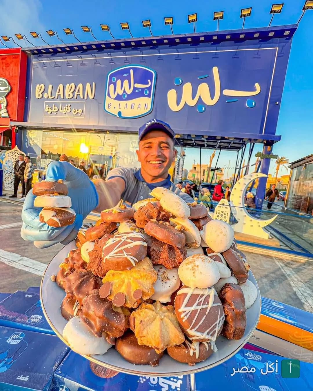 موظف في بـ لبن عين شمس يقدم طبقاً كبيراً من البسكويت المغطى بالشوكولاتة أمام واجهة الفرع.