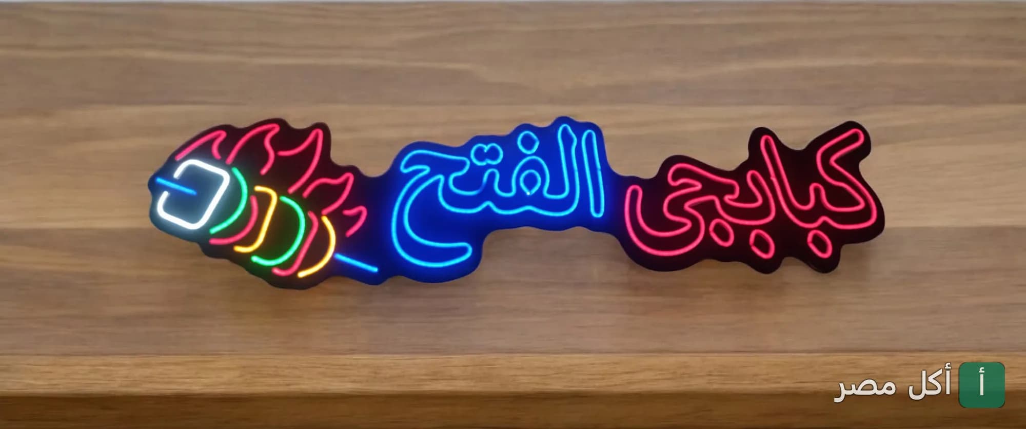 لوحة نيون مضيئة باسم كبابجي الفتح مع رسم سيخ مشويات ملون على خلفية خشبية.