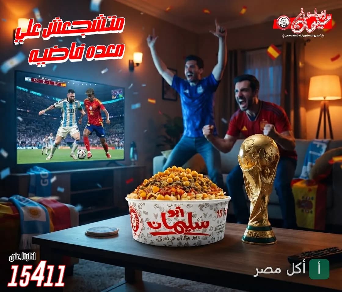 علبة مكرونة من أبو سليمان على طاولة أمام شاشة تعرض مباراة كرة قدم مع مجسم لكأس العالم.