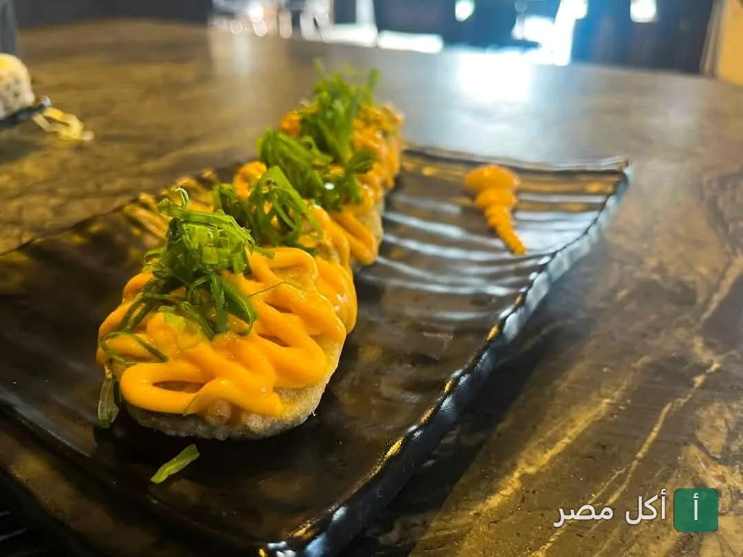 قطع سوشي مغطاة بصوص أورانج وشرائح البصل الأخضر على طبق أسود في مطعم سوشي هوم بالخليفة.