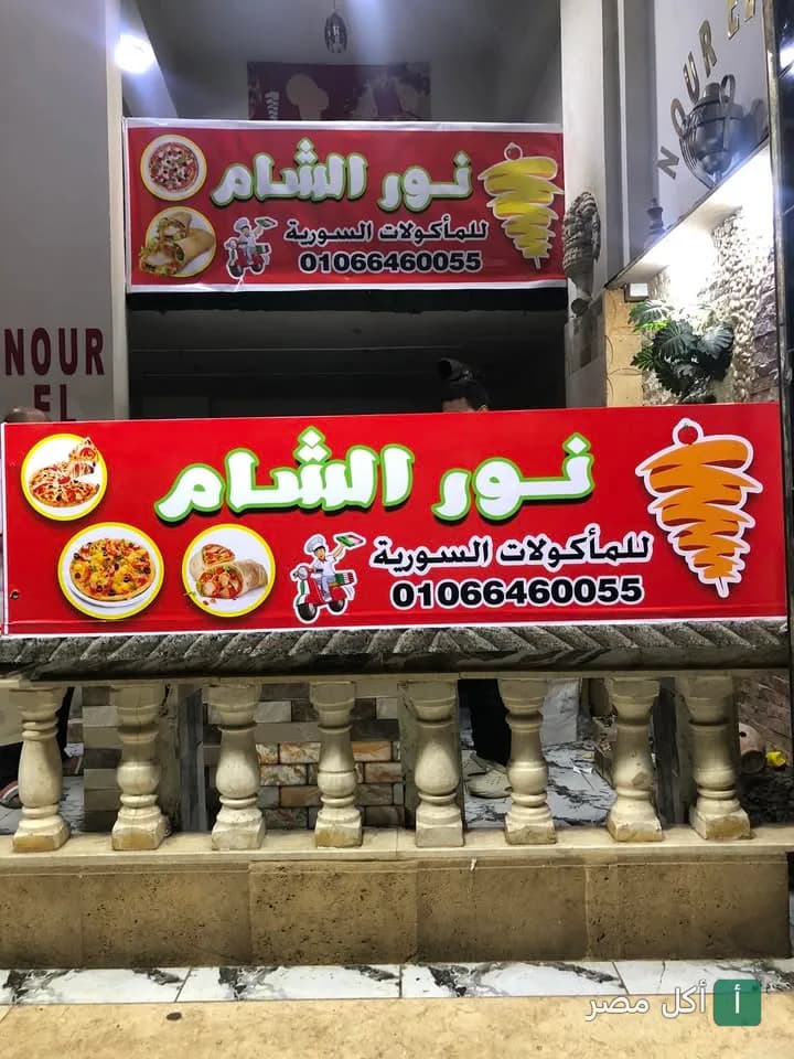 واجهة مطعم نور الشام للمأكولات السورية في الخليفة مع لافتة حمراء تعرض صور البيتزا والشاورما.