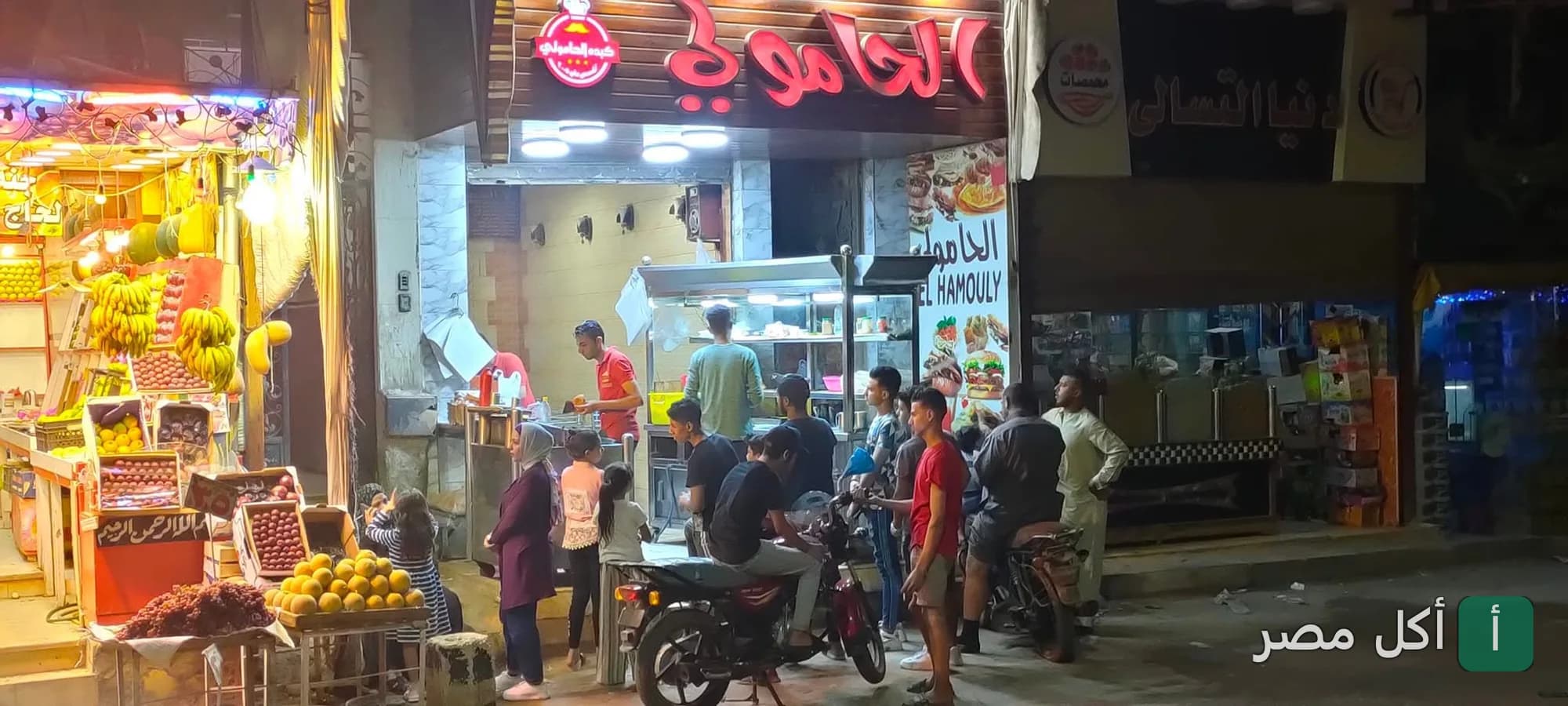 واجهة مطعم كبدة الحامولي في الخليفة ليلاً مع زبائن ينتظرون الطلبات أمام المحل.