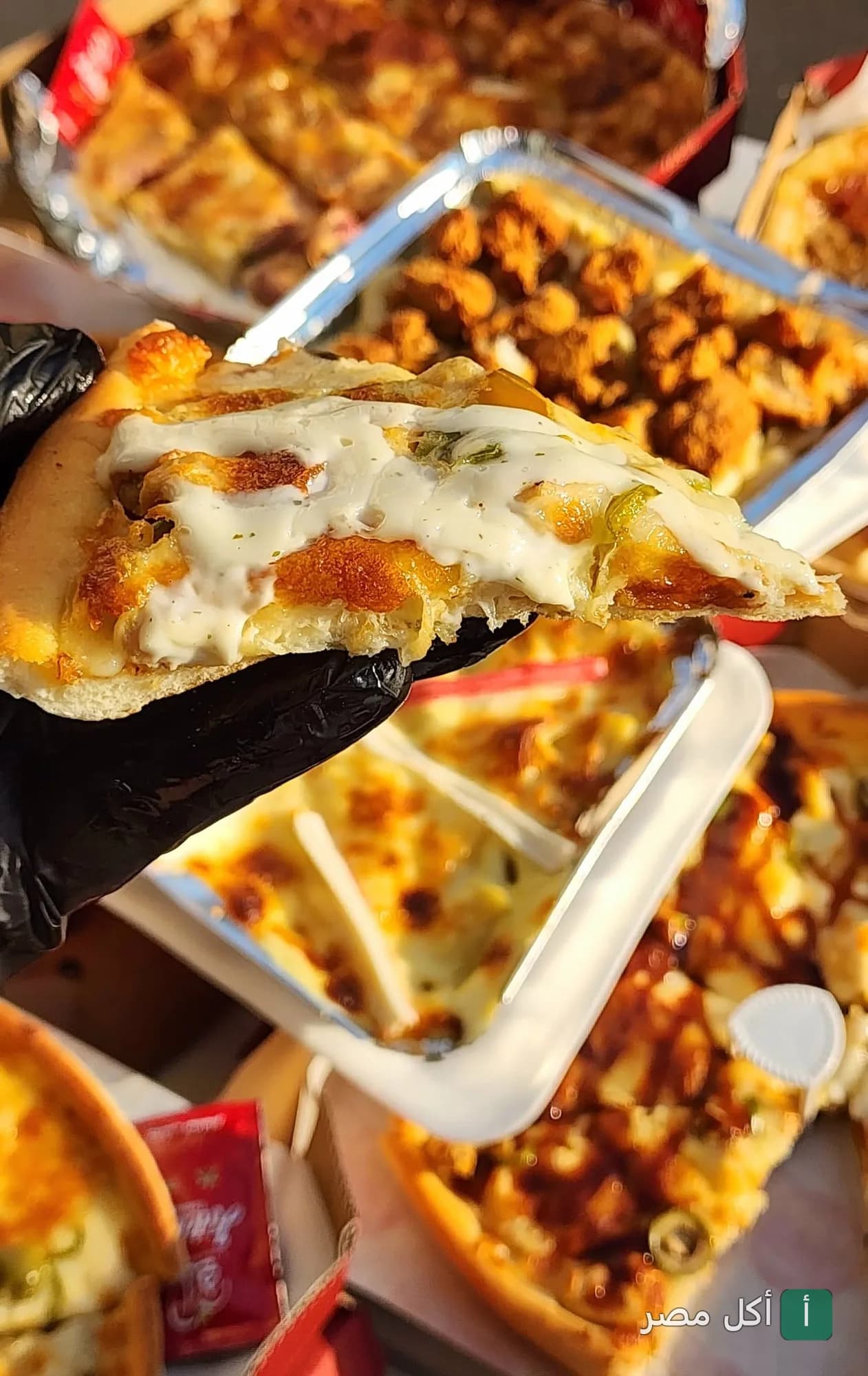قطعة بيتزا غنية بالجبنة والصوص من مطعم تيبستي الكوربه، مع مجموعة متنوعة من الوجبات السريعة في الخلفية.