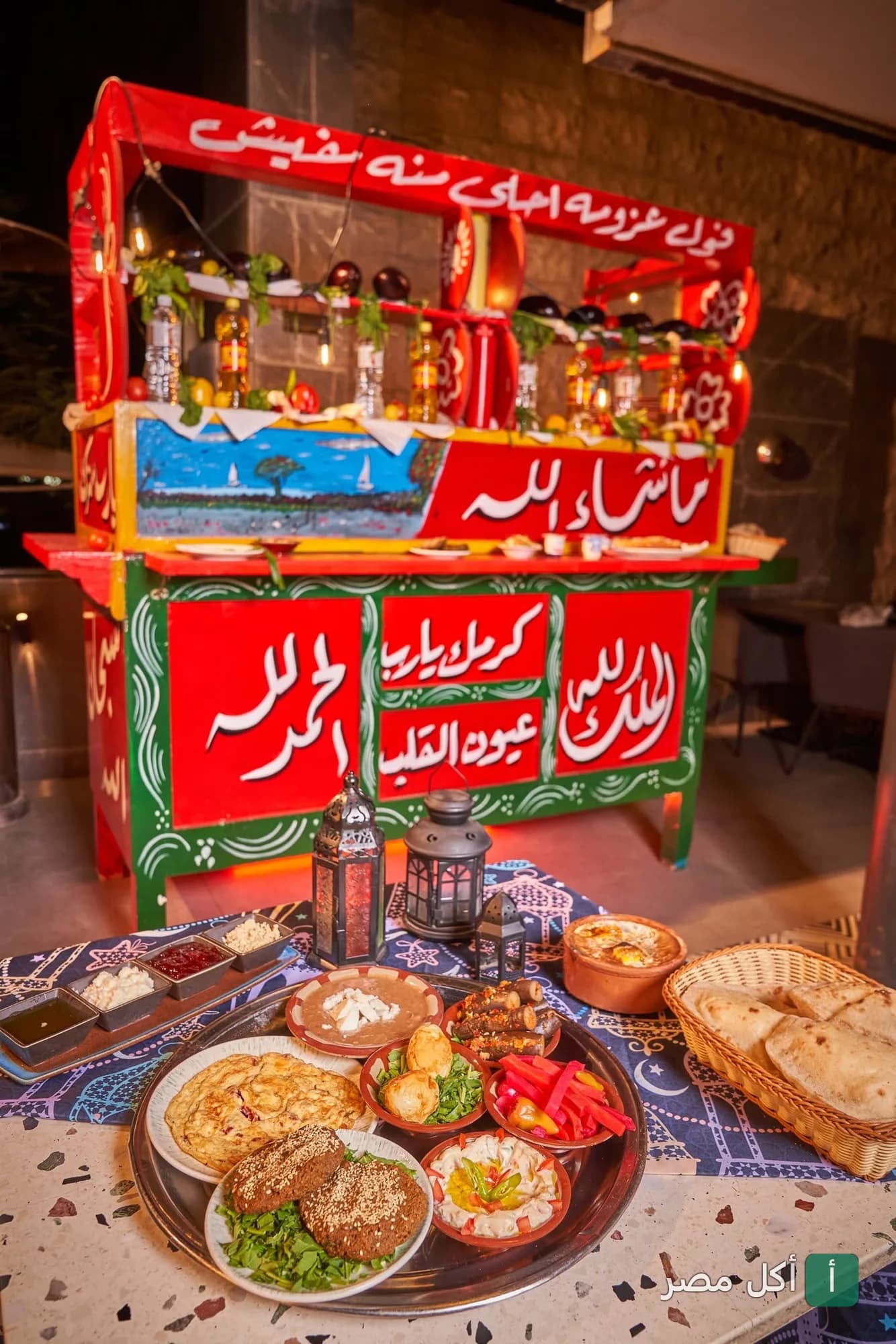 عربة فول تقليدية ملونة في مطعم عزومة، مع سفرة إفطار مصرية متنوعة تشمل الفول والطعمية والخبز البلدي.