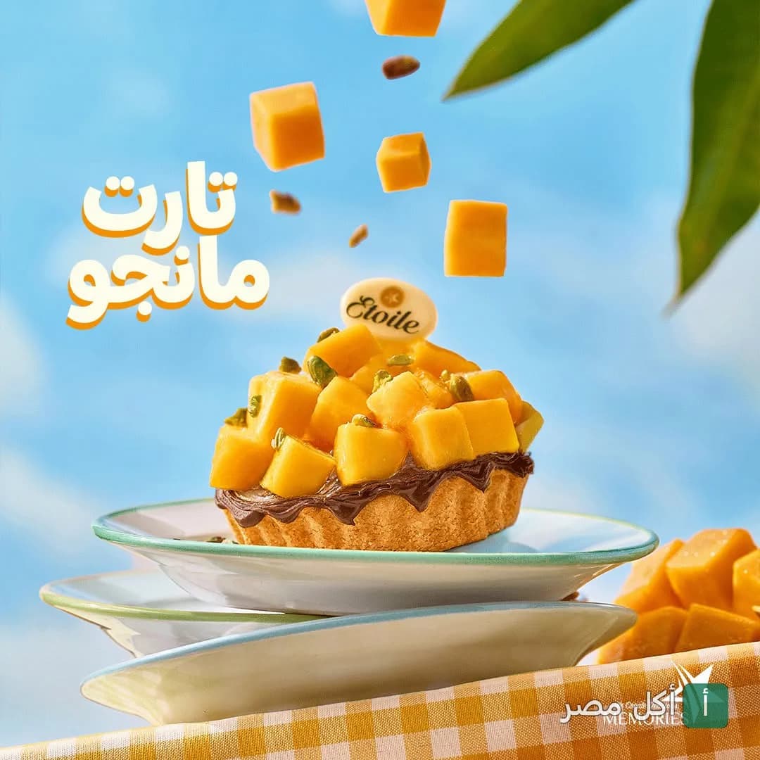 تارت مانجو من إيتوال فيصل، مزين بقطع المانجو الطازجة والفستق على خلفية سماوية.