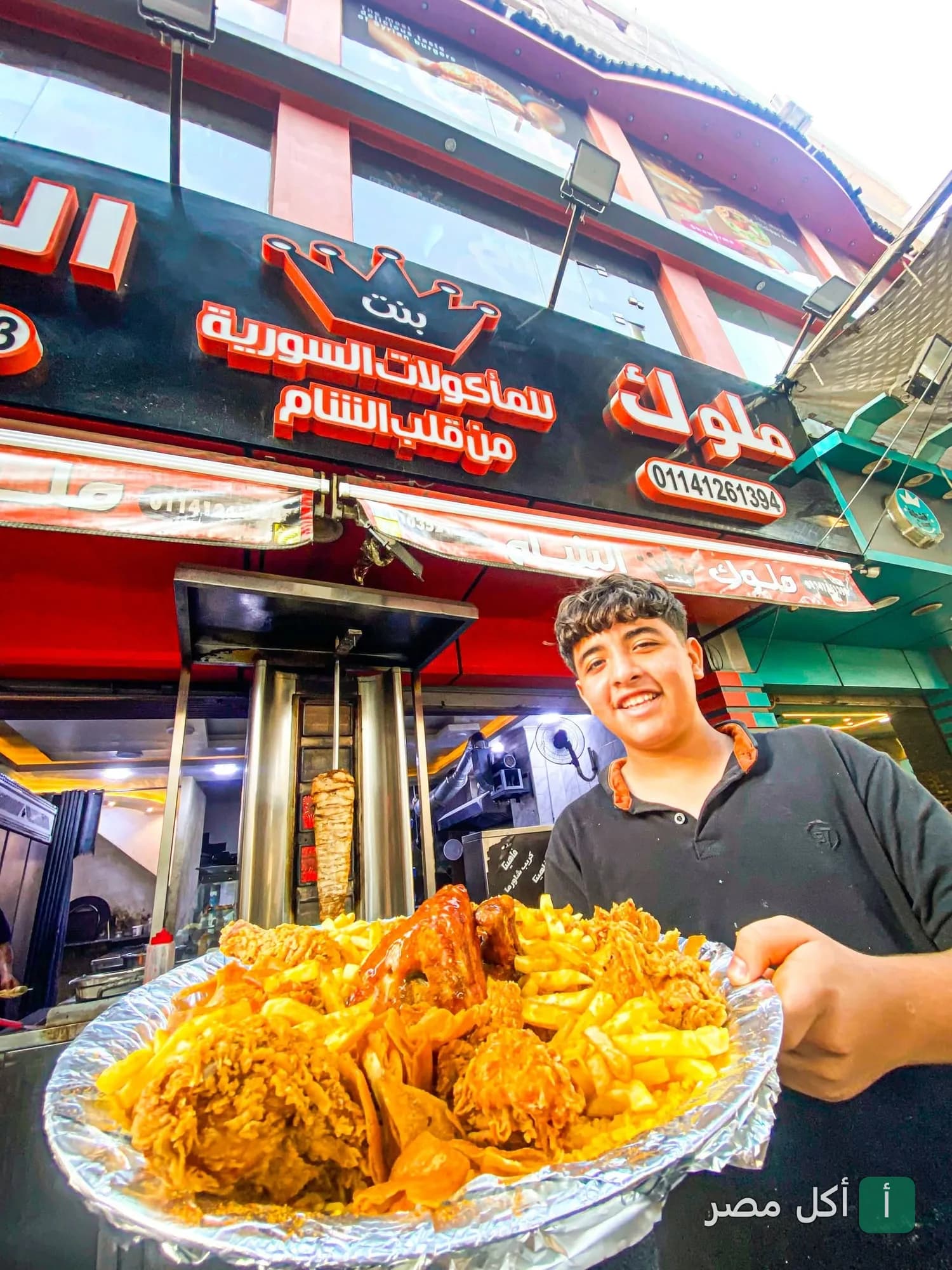 موظف في مطعم ملوك بنت الشام ببولاق الدكرور يقدم طبق دجاج مقلي مع بطاطس محمرة أمام واجهة المطعم.