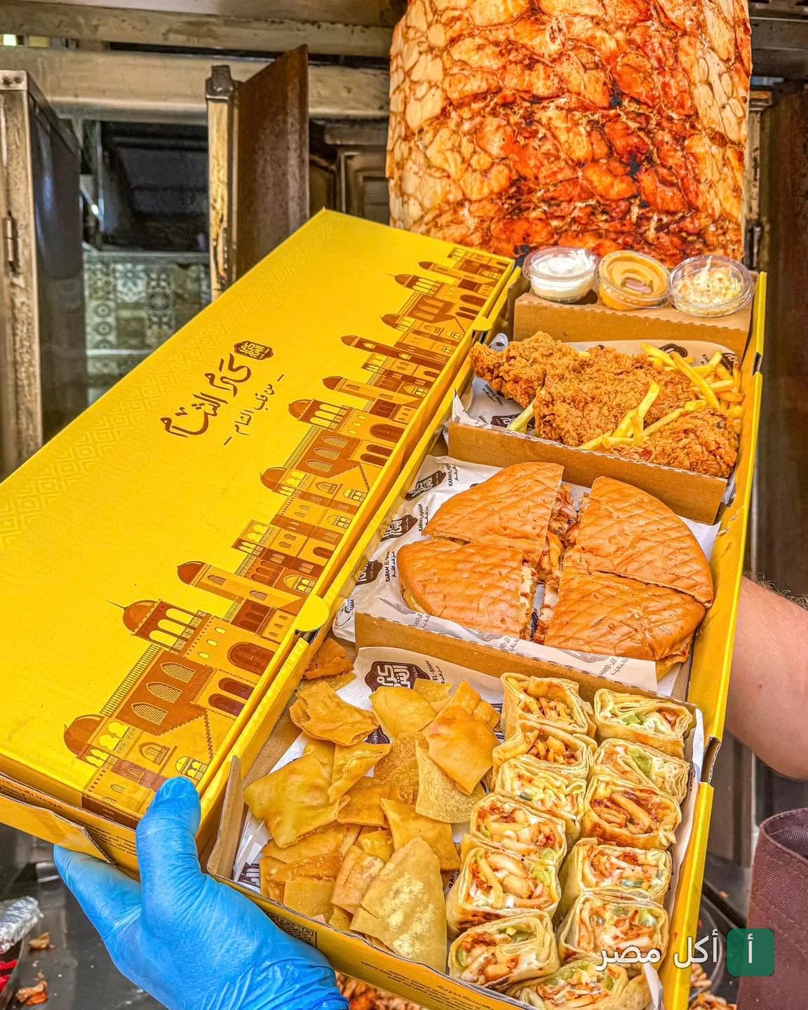 صندوق وجبات كرم الشام يضم شاورما سورية مقطعة، دجاج مقلي، بطاطس مقلية ومقبلات أمام سيخ الشاورما.