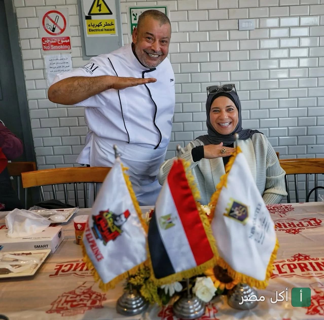 الشيف ناصر البرنس يبتسم بجانب زائرة في مطعم البرنس بإمبابة، مع أعلام صغيرة على طاولة الطعام.