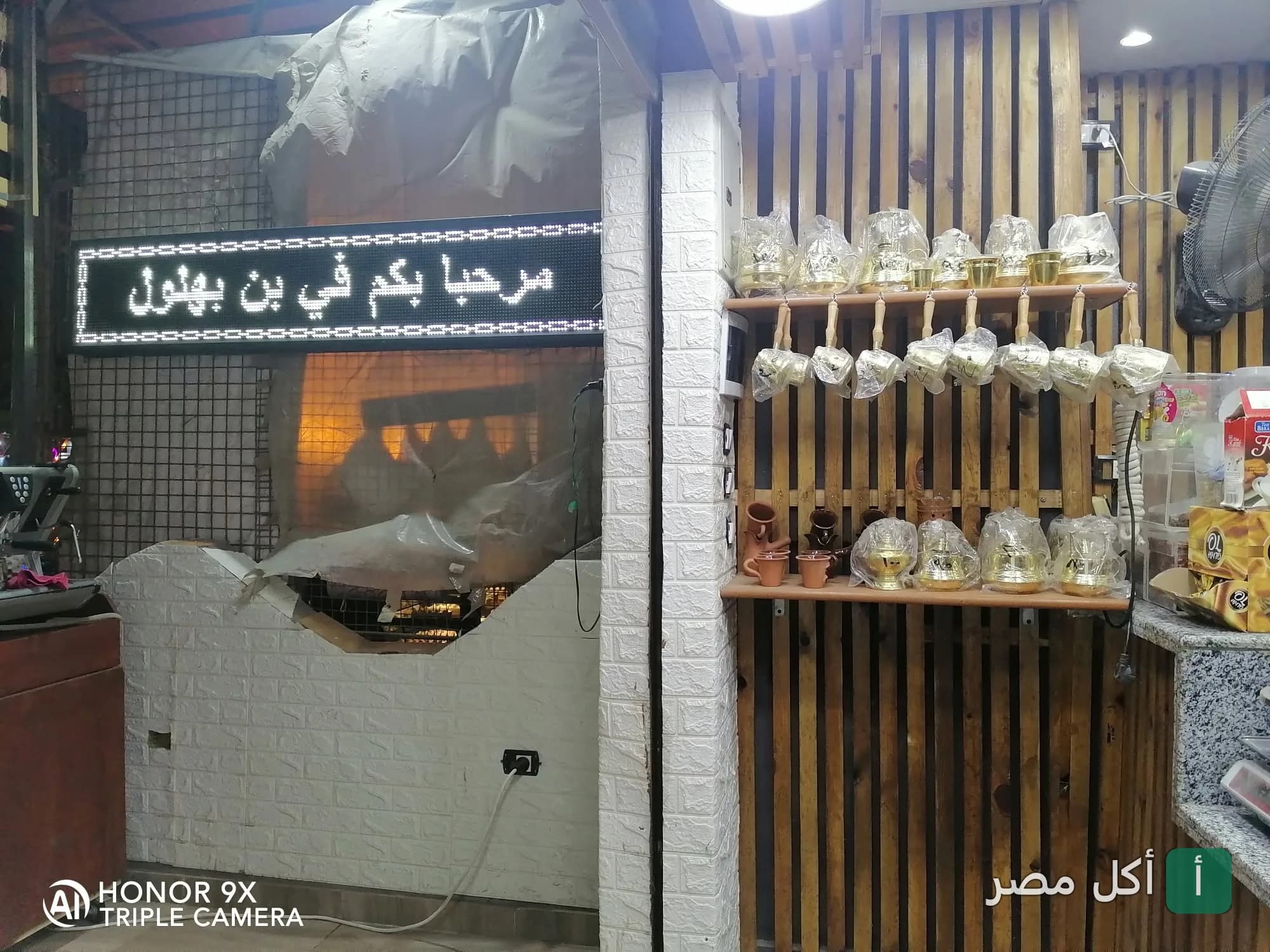 ديكور داخلي في بهلول كافيه بالمنيرة في إمبابة يظهر أرفف خشبية عليها أواني نحاسية وشاشة إلكترونية ترحب بالزبائن.