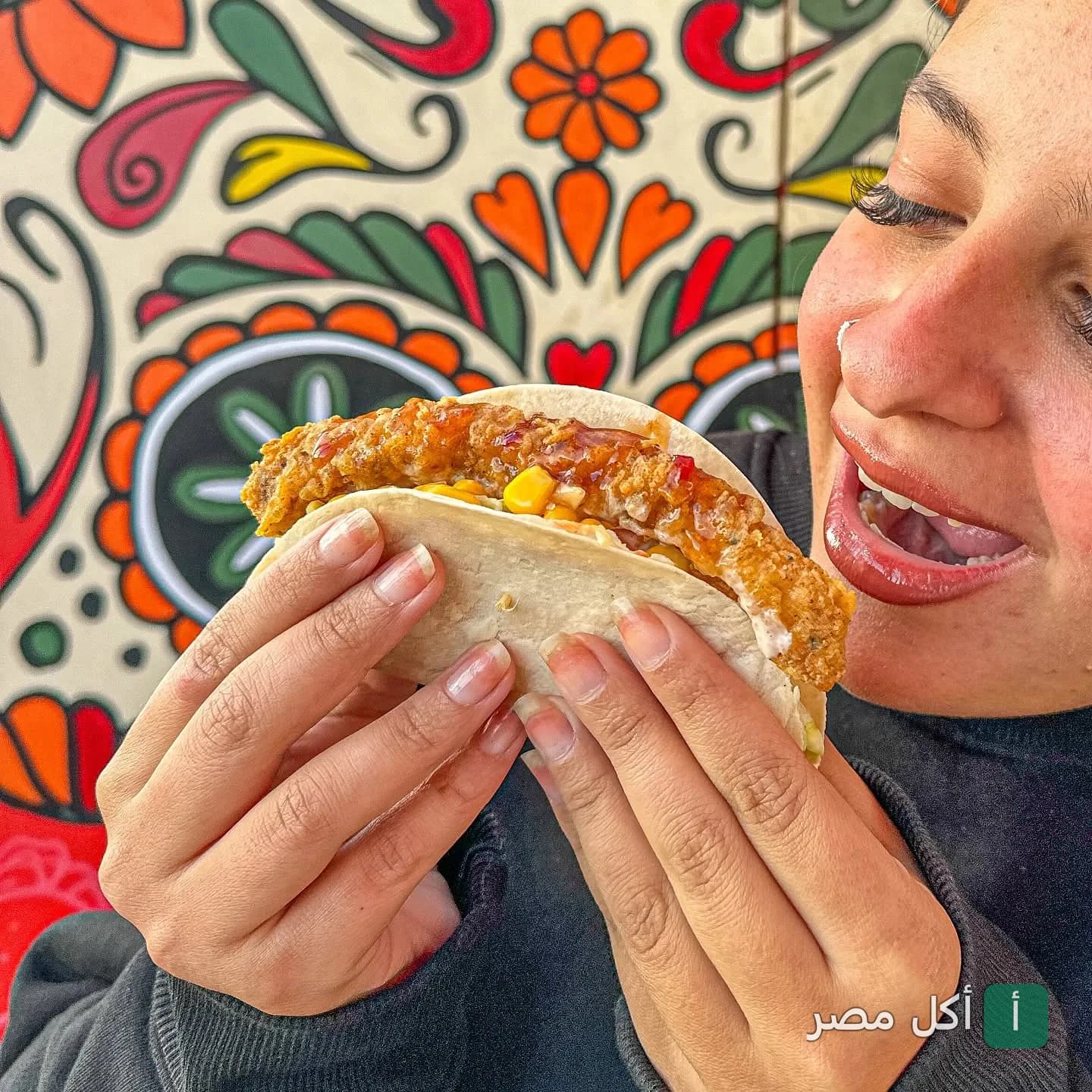 شخص يتناول وجبة سريعة في مطعم هيرمانوس بالشيخ زايد أمام حائط مزين بنقوش ملونة.