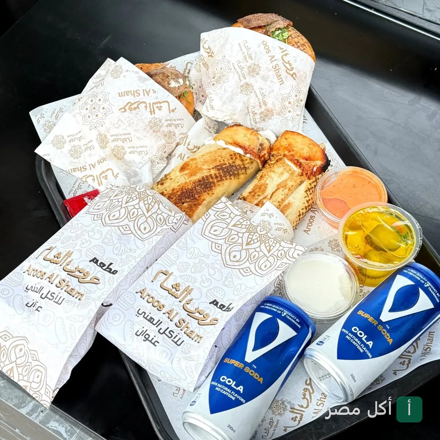 صينية تحتوي على ساندوتشات شاورما مغلفة بورق مطبوع عليه شعار مطعم عروس الشام في الشيخ زايد مع علب صوص ومشروبات غازية.