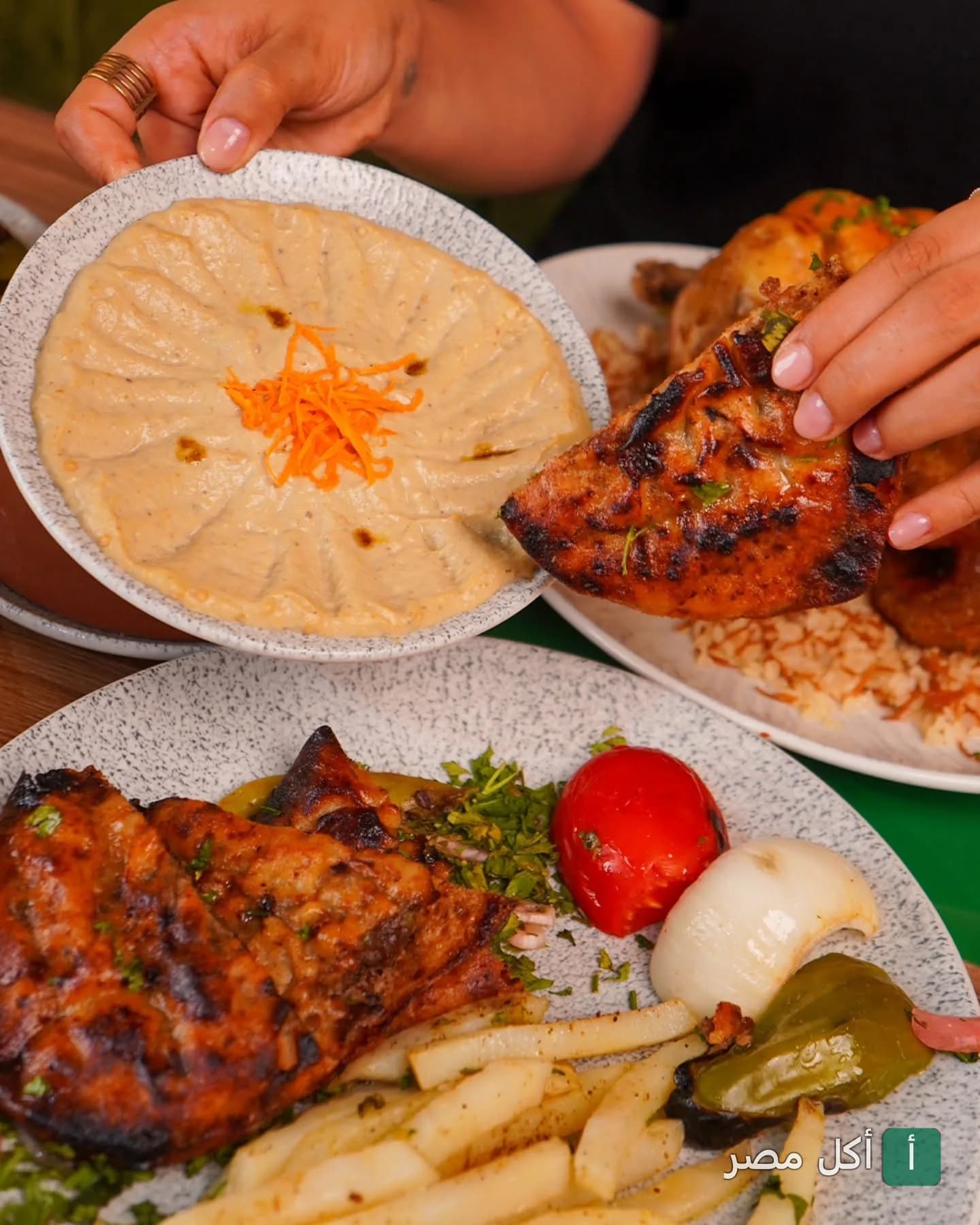 شخص يغمس قطعة دجاج مشوي في طبق طحينة مع أطباق جانبية من الدجاج المشوي والبطاطس في مطعم محشي ماما بالوراق.