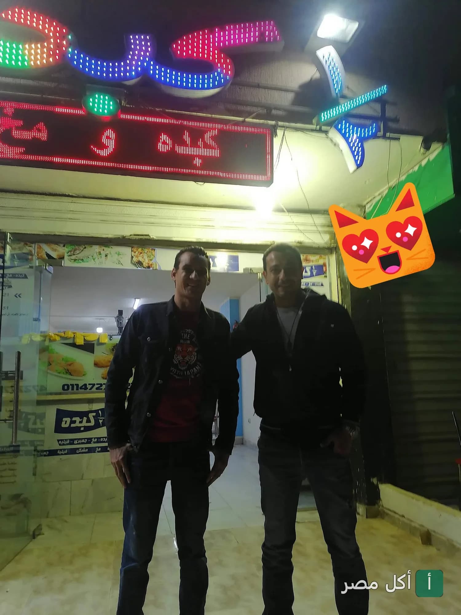 شخصان يقفان أمام واجهة مطعم نص كبده في الأهرام ليلاً مع لافتة نيون مضيئة.