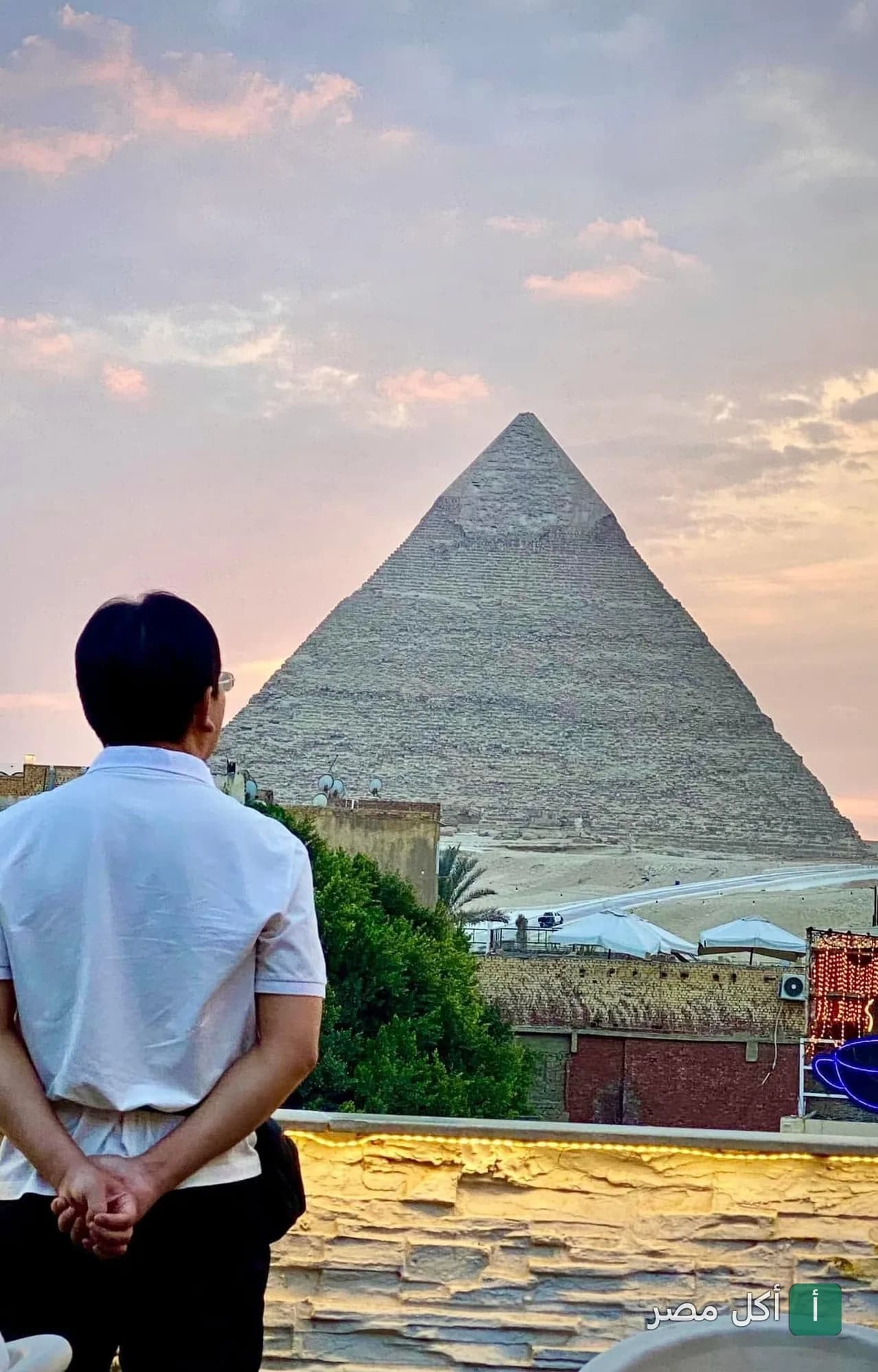 شخص يقف في تراس روكان كافيه بنزلة السمان ويشاهد أهرامات الجيزة وقت الغروب.