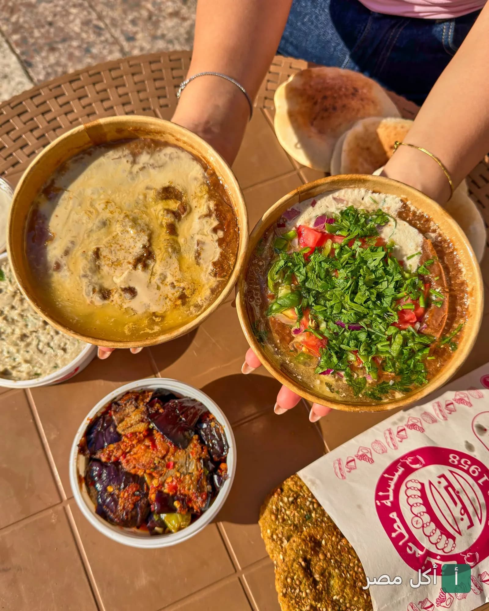 شخص يحمل أطباق فول شرقي متنوعة مع باذنجان مقلي وخبز طازج من مطاعم نجف بالصف.