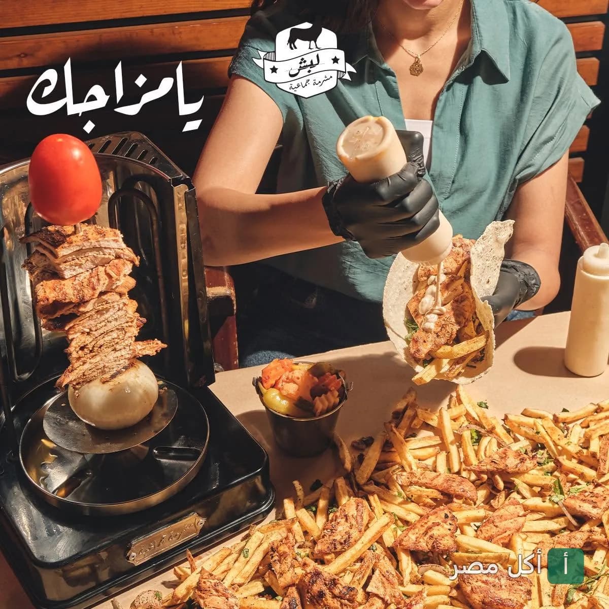 شخص يضيف الصوص لساندوتش شاورما بجانب سيخ شاورما صغير وكمية كبيرة من البطاطس في مطعم لبش بالإسكندرية.