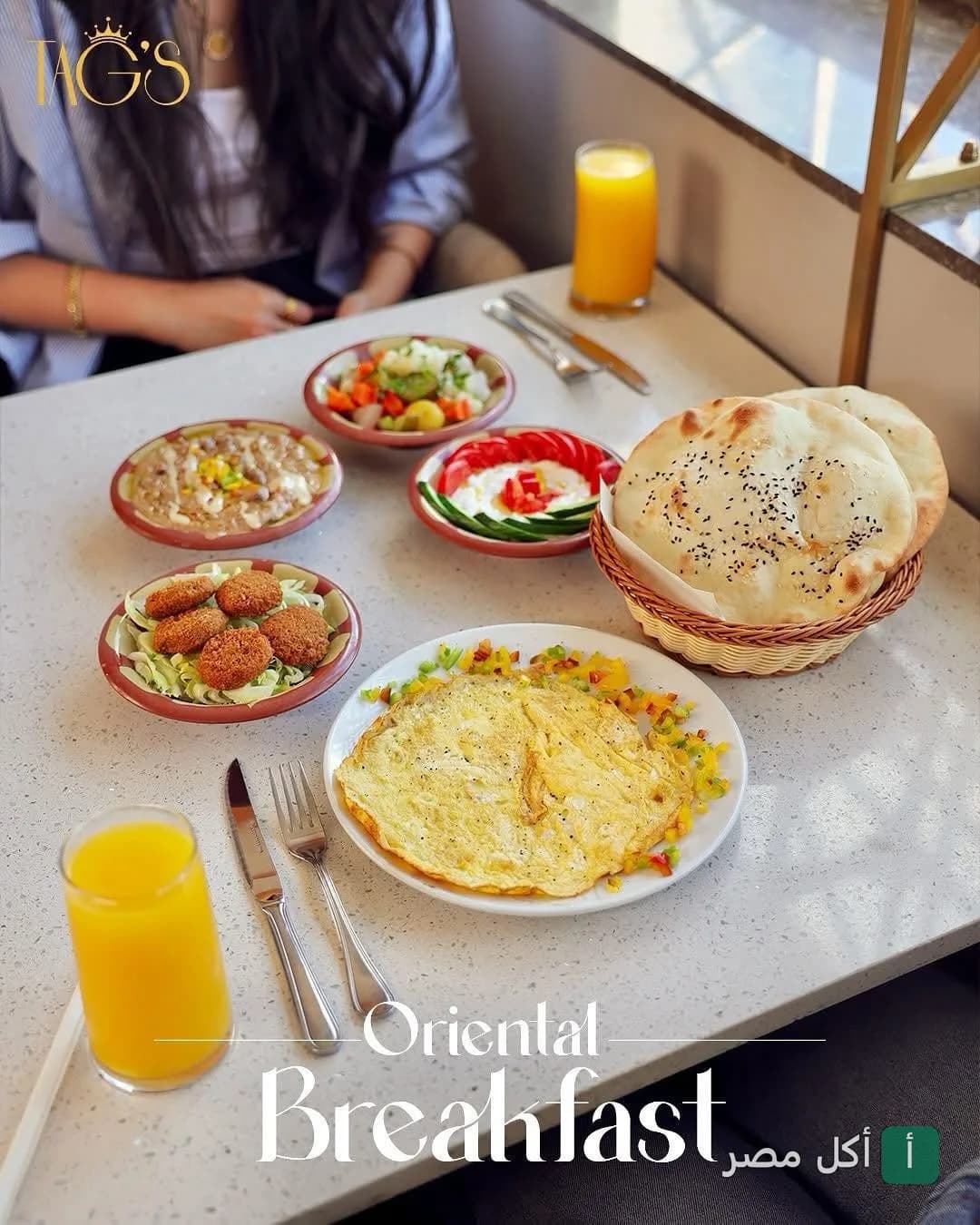 سفرة فطار شرقي متنوعة في مطعم تاجز بسيدي جابر، تشمل أومليت، فول، طعمية، وجبنة بالطماطم مع خبز طازج وعصير برتقال.