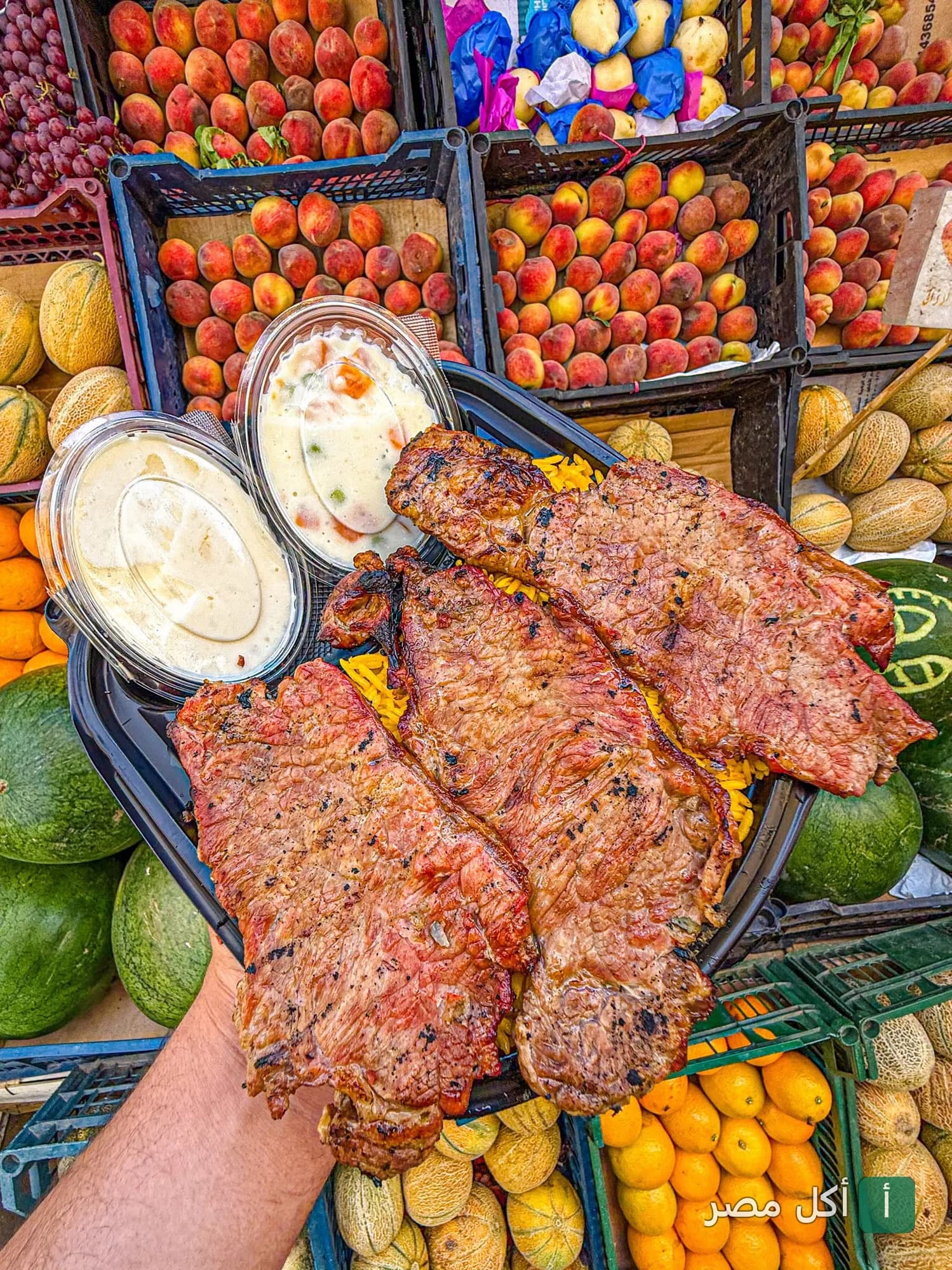 وجبة مشويات من مطعم الجمّال في مينا البصل، تتكون من قطع لحم مشوية مع أرز وسلطات، محاطة بصناديق فاكهة طازجة.