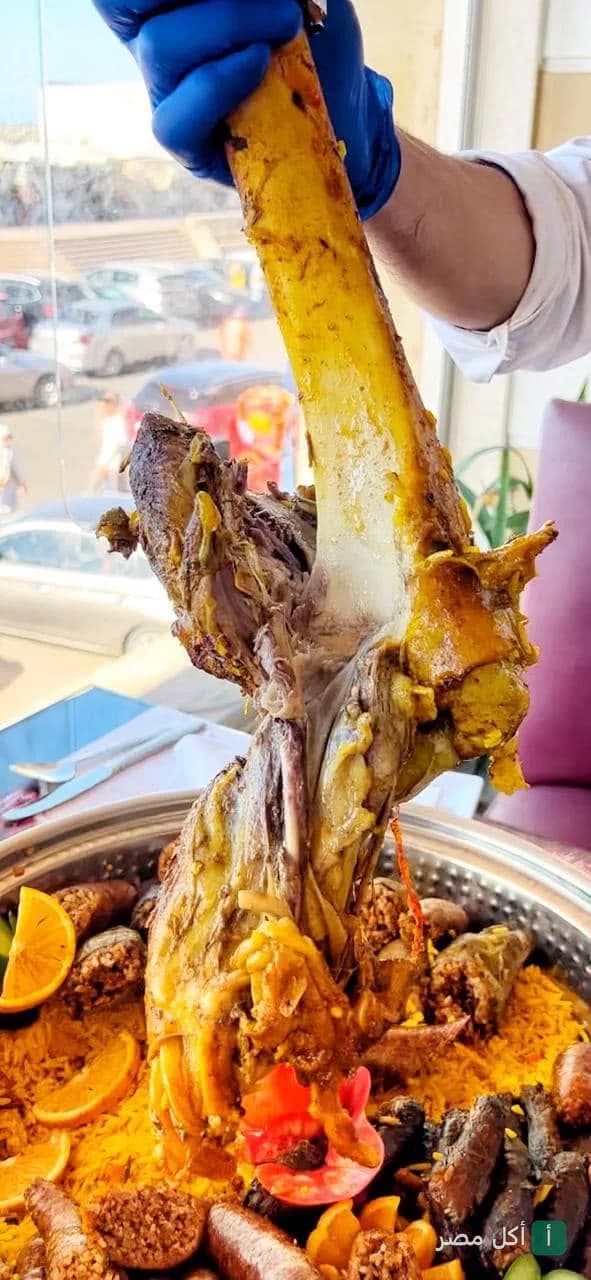 يد ترتدي قفاز أزرق ترفع قطعة كبيرة من لحم الموزة الضاني فوق طبق أرز بالمكسرات ومحشي في مطعم قلعة المشويات بالإسكندرية.