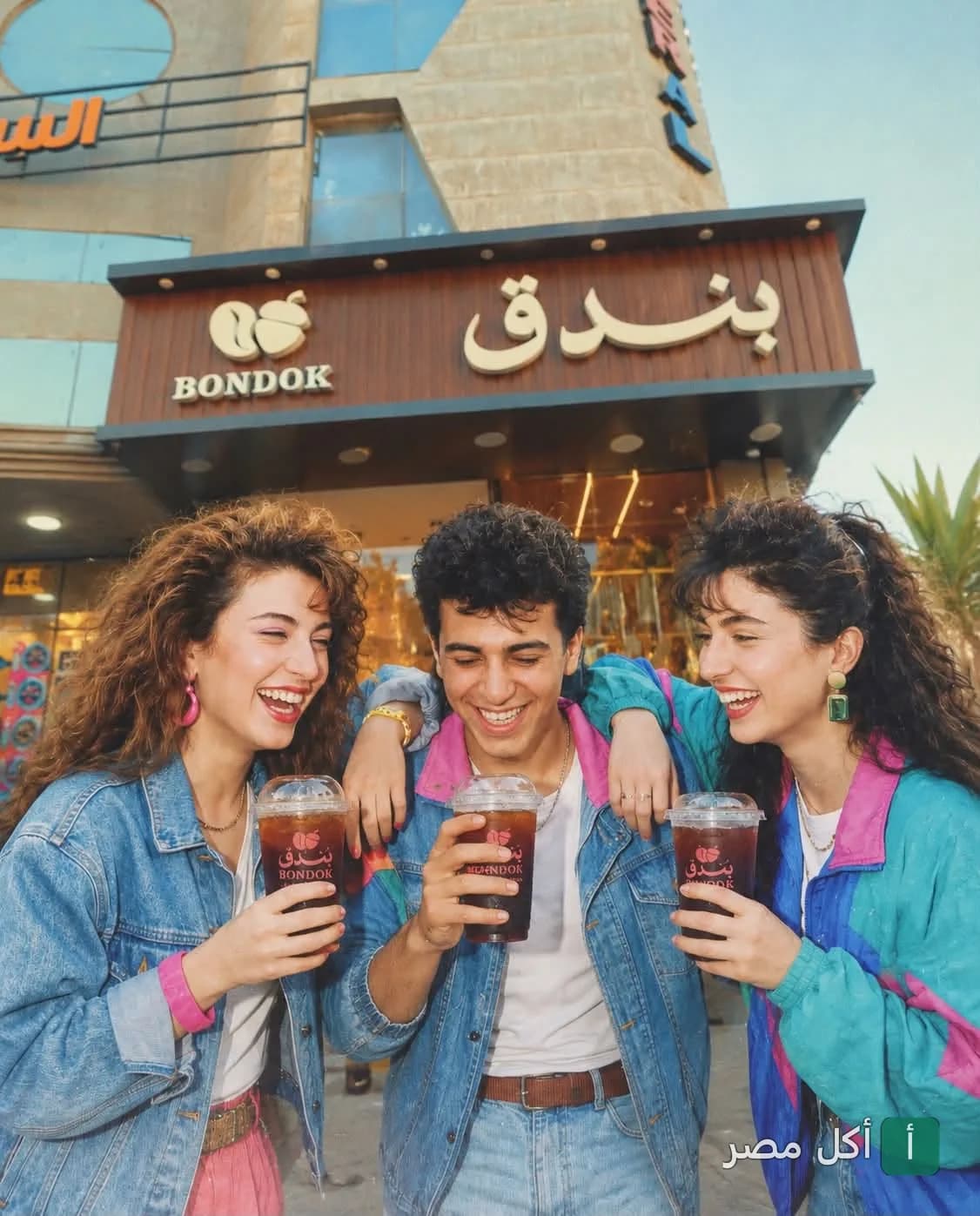 ثلاثة أصدقاء يضحكون ويحملون مشروبات باردة أمام واجهة كافيه بندق في مينا البصل بالإسكندرية.